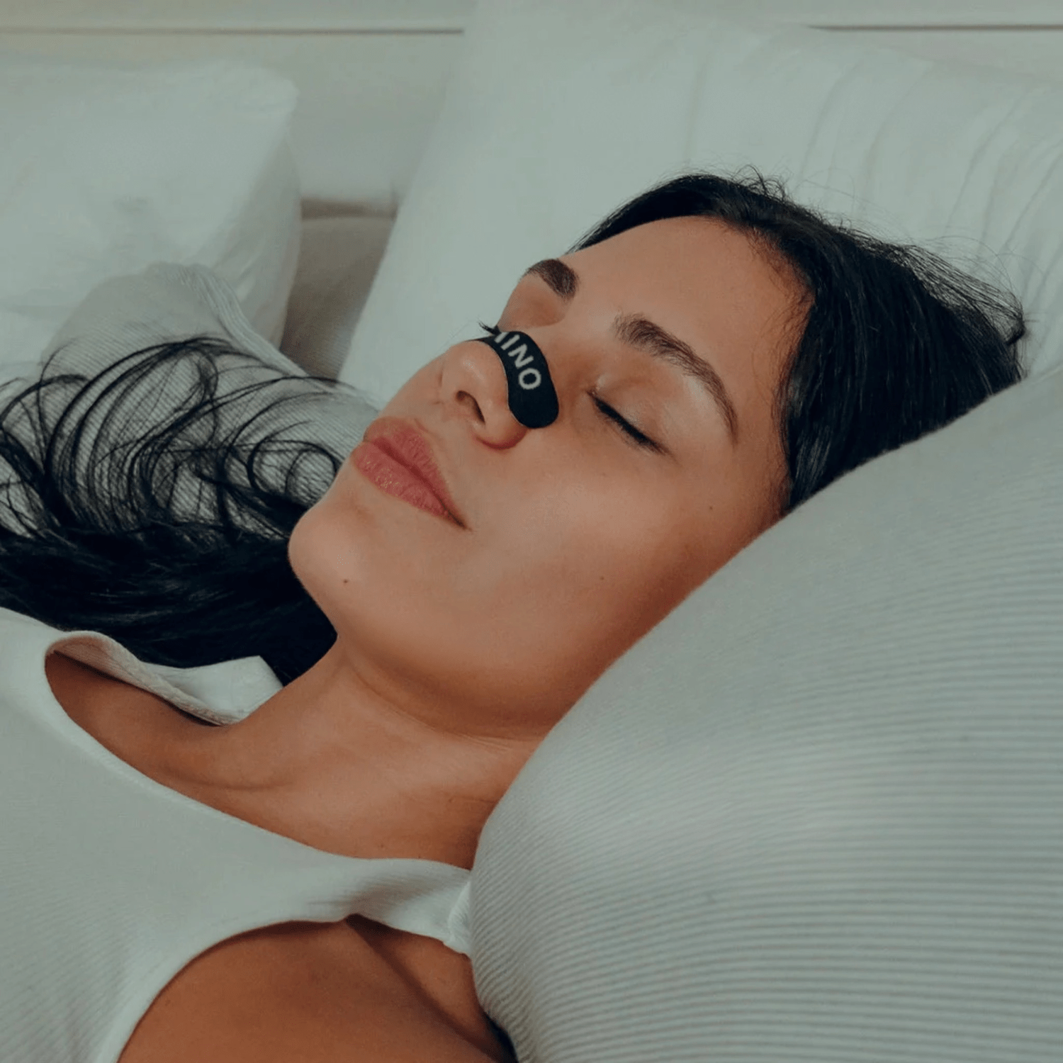Woman lying in bed with anose strip on, wearing a white shirt.